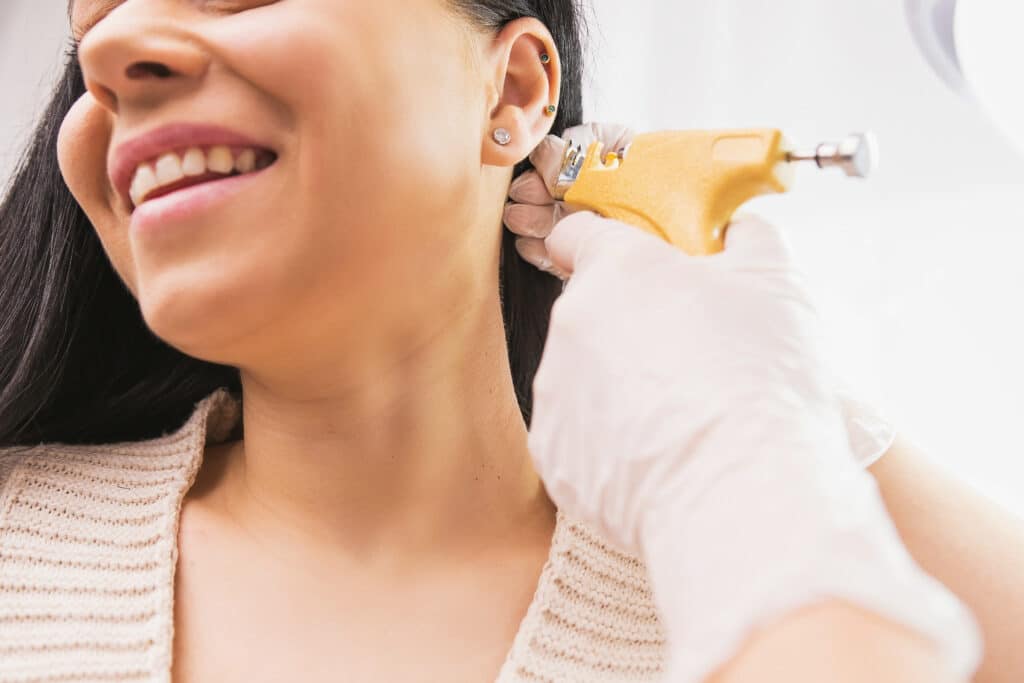 Cropped photo of a smiling woman patient getting ear piercing procedure by a specialist in medical gloves in the cosmetologist cabinet. Smiling woman getting ear piercing procedure by a specialist